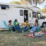 Modern travel trailer toy hauler with an extended ramp patio, an ATV, and a couple enjoying the scenic outdoor adventure by a lake.